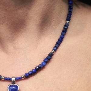 Op de afbeelding: Een donkerblauwe kralenketting met een hartvormige hanger. De hanger is van zilverkleurig metaal met een donkerblauwe steen in de vorm van een hart. De ketting is gemaakt van kleine, ronde, donkerblauwe kralen en zilverkleurige metalen kralen.