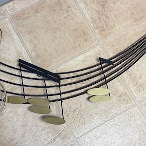 Könnte beinhalten: Dekorative Notenlinien aus Metall mit Violinschlüssel und Noten. Die Notenlinien sind schwarz mit goldfarbenen Noten. Der Violinschlüssel ist silberfarben. Zur Wandmontage.