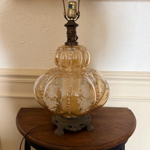 May include: A vintage amber glass table lamp with a floral design etched into the glass. The lamp has a brass base and a decorative metal base. The lamp is sitting on a small, round, dark wood table.
