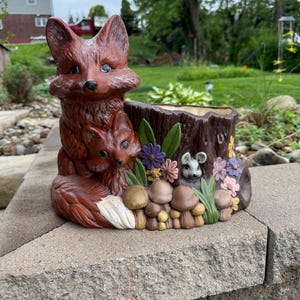 May include: A brown ceramic planter featuring a fox family, a mouse, and mushrooms. The planter is shaped like a tree stump with colorful flowers and green leaves. The fox has a white tail and is sitting next to its baby.