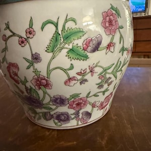 May include: A white ceramic planter with a floral design. The planter features pink, purple, and green flowers and leaves. The rim has a green and purple border. The planter is likely used for indoor or outdoor plants.