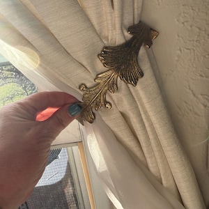 May include: A decorative gold-colored curtain tieback in a leaf design. The tieback is holding a cream-colored curtain. The curtain is made of a textured fabric. The tieback is made of metal.
