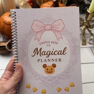 May include: A spiral-bound notebook with a light pink cover featuring a large pink bow, a decorative frame, and the text Castle Keys Co. Magical Planner. Below the text is a pumpkin icon with mouse ears, and candy corn illustrations.