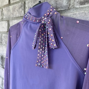Op de afbeelding: Een lavendelkleurig paardrijshirt met een hoge kraag en lange mouwen. De kraag en strik zijn versierd met iriserende strass-steentjes. Mesh panelen bevinden zich op de bovenarmen en schouders. Het shirt hangt aan een zwarte hanger.