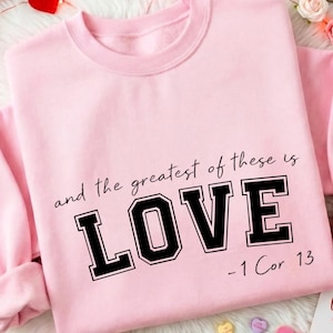 Puede incluir: Sudadera rosa claro con la frase "and the greatest of these is LOVE -1 Cor 13" en negro. La imagen incluye decoraciones de San Valentín como corazones, flores y una cinta roja.