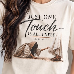 Puede incluir: Una camiseta de manga corta de color crema con un estampado gráfico de una persona extendiendo la mano para tocar una túnica. El texto "JUST ONE Touch IS ALL I NEED - MARK 5:28 -" aparece en letras negras sobre la ilustración.