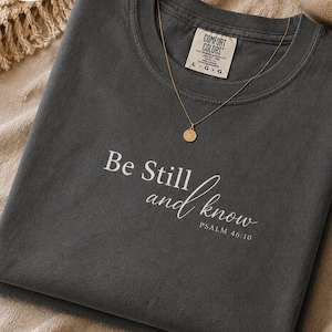 May include: A pepper-colored t-shirt featuring the white printed text Be Still and know PSALM 46:10. The shirt has a Comfort Colors brand label on the inner neckline indicating a size large and a gold-toned necklace with a circular pendant.