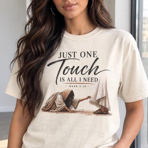 May include: A cream-colored short-sleeve t-shirt featuring a graphic illustration of a person reaching out to touch the hem of a robe. The text reads: JUST ONE Touch IS ALL I NEED - MARK 5:28 - in black and brown lettering on the front of the shirt.