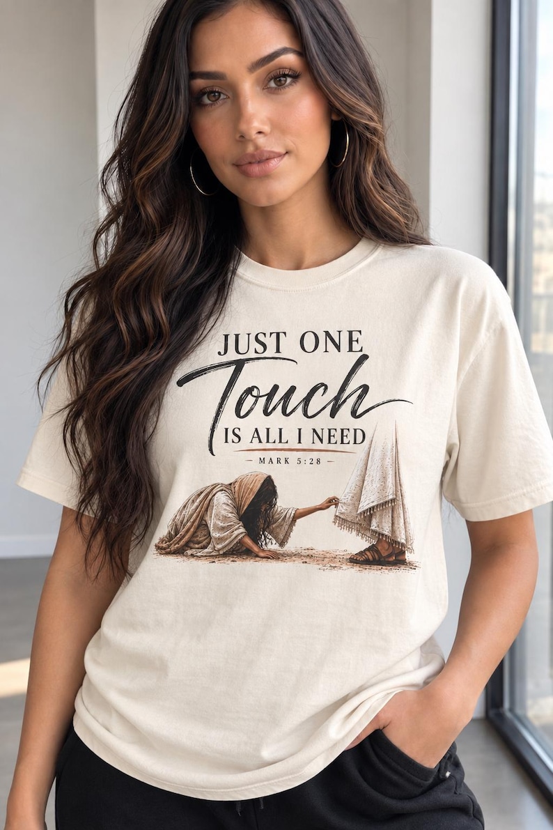 May include: A cream-colored short-sleeve t-shirt featuring a graphic illustration of a person reaching out to touch the hem of a robe. The text reads: JUST ONE Touch IS ALL I NEED - MARK 5:28 - in black and brown lettering on the front of the shirt.
