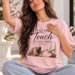 May include: A light pink t-shirt featuring a graphic illustration of a person reaching out to touch a robe, with the text "JUST ONE Touch IS ALL I NEED" and "MARK 5:28" printed in black, paired with light blue denim jeans and white sneakers.
