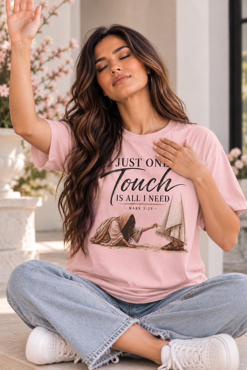 May include: A light pink t-shirt featuring a graphic illustration of a person reaching out to touch a robe, with the text "JUST ONE Touch IS ALL I NEED" and "MARK 5:28" printed in black, paired with light blue denim jeans and white sneakers.