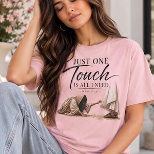 May include: A light pink short-sleeve t-shirt featuring the black text "JUST ONE Touch IS ALL I NEED - MARK 5:28 -" above a graphic illustration of a person reaching out to touch the hem of a robe worn by another figure.