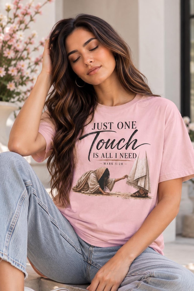 May include: A light pink short-sleeve t-shirt featuring the black text "JUST ONE Touch IS ALL I NEED - MARK 5:28 -" above a graphic illustration of a person reaching out to touch the hem of a robe worn by another figure.