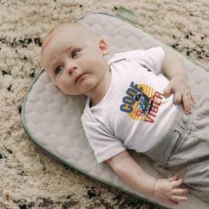 Può includere: Un body bianco per neonati con la scritta "CODE VIBER" in blu e rosso. La grafica presenta un astronauta e un motivo floreale su uno sfondo giallo e arancione. Il bambino è sdraiato su un tappetino imbottito grigio.