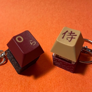 May include: Two keychains with keycaps. One keycap is burgundy with the number 5 and the letter O. The other keycap is tan with a Japanese character. Both keycaps are attached to keyrings.
