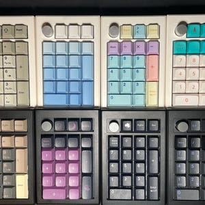 May include: Eight mechanical number pads arranged in two rows of four. The top row features white cases with various colored keycaps, while the bottom row has black cases with dark, purple, and grey keycaps. Each pad includes a circular knob.