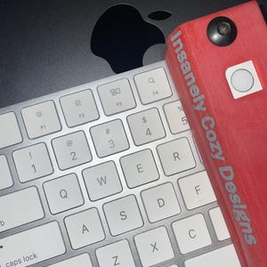 May include: A silver and white computer keyboard with a red rectangular object that has the text "Insanely Cozy Designs". The keyboard is on a black surface with an Apple logo.