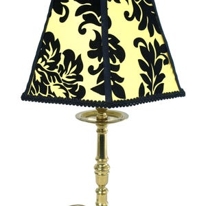 May include: A brass table lamp with a yellow lampshade featuring a black floral pattern. The lampshade is trimmed with black fabric and a decorative edge. A clear, round finial tops the shade.
