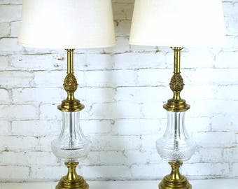 May include: Pair of vintage table lamps with cream-colored fabric shades. The lamps feature a clear glass body with a diamond pattern, brass accents, and a pineapple-shaped finial. The lamps are wired and ready to use.