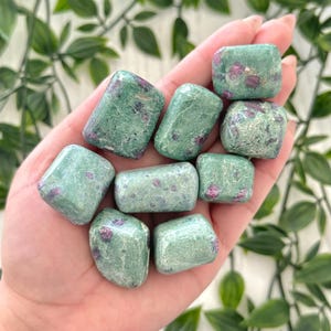 May include: A handful of green tumbled stones with red flecks. The stones are smooth and rounded, varying in size. The background shows green leaves. The stones are likely measured in centimetres.