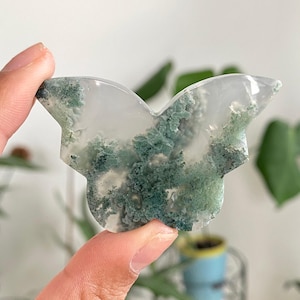 Peut inclure: Une sculpture en pierre en forme de papillon avec un motif blanc et vert translucide. Les inclusions vertes ressemblent à de la mousse ou du feuillage, créant un design naturaliste. La pierre est tenue par les doigts d'une personne.