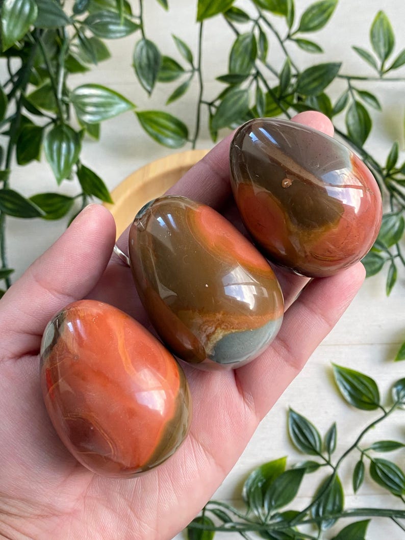 May include: Three polished gemstone eggs in shades of brown, orange, and gray are held in a hand. The eggs have a smooth, glossy finish and a natural, marbled appearance. The background includes green foliage.