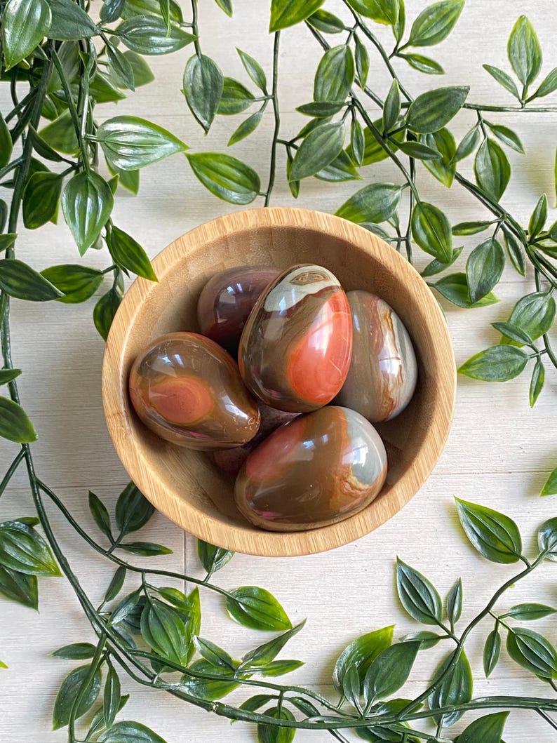 May include: A wooden bowl filled with polished, egg-shaped stones. The stones display a mix of colors, including brown, red, and gray, with a glossy finish. Green leafy vines surround the bowl, creating a natural aesthetic.