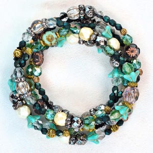 Könnte beinhalten: Ein mehrreihiges Armband mit türkisfarbenen, grünen, goldenen und transparenten Perlen. Die Perlen umfassen facettierte Kristalle, florale Akzente und perlenartige Perlen. Das Armband ist kreisförmig auf weißem Hintergrund angeordnet.
