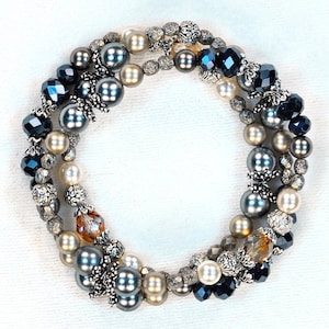 May include: A multi-strand wrap bracelet featuring a mix of dark grey, cream, and white pearls, faceted dark blue crystal beads, and textured silver-toned metal spacers. The design incorporates clear amber-toned crystals and ornate silver bead caps.
