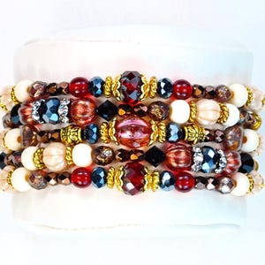 Puede incluir: Pulseras apiladas con cuentas de colores variados. Las pulseras incluyen cuentas rojas, negras, doradas y color crema. Algunas cuentas tienen un diseño facetado, y otras son redondas u ovaladas. Las pulseras se exhiben sobre una superficie blanca.