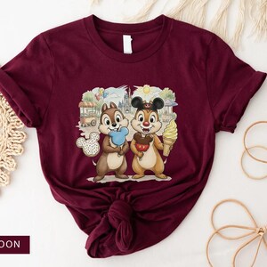 May include: Maroon t-shirt with a graphic of two cartoon chipmunks, each holding a treat against a theme park background. One chipmunk has blue candy floss, the other an ice cream cone. The tee shirt is a solid maroon colour.