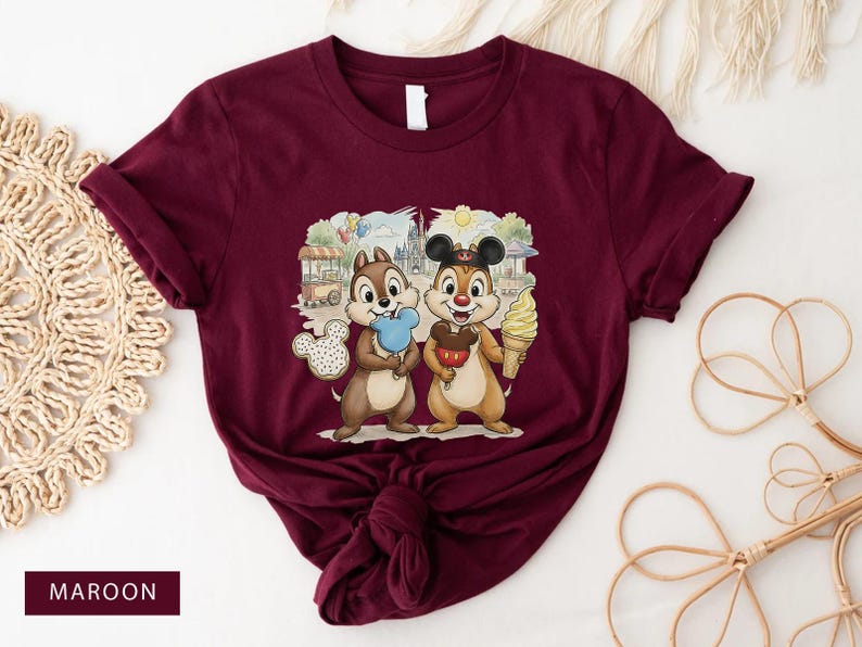 May include: Maroon t-shirt with a graphic of two cartoon chipmunks, each holding a treat against a theme park background. One chipmunk has blue candy floss, the other an ice cream cone. The tee shirt is a solid maroon colour.