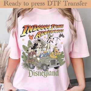 May include: A light pink t-shirt featuring a graphic print of Mickey Mouse, Minnie Mouse, Donald Duck, Goofy, and Pluto in explorer gear riding in a jeep. The text reads Indiana Jones Adventures and Disneyland. Ready to press DTF transfer.