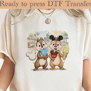 May include: Cream-coloured t-shirt with a DTF transfer of Chip and Dale. Chip holds a blue popsicle, and Dale holds a Mickey Mouse ice cream. The background shows a castle, food cart, and balloons.