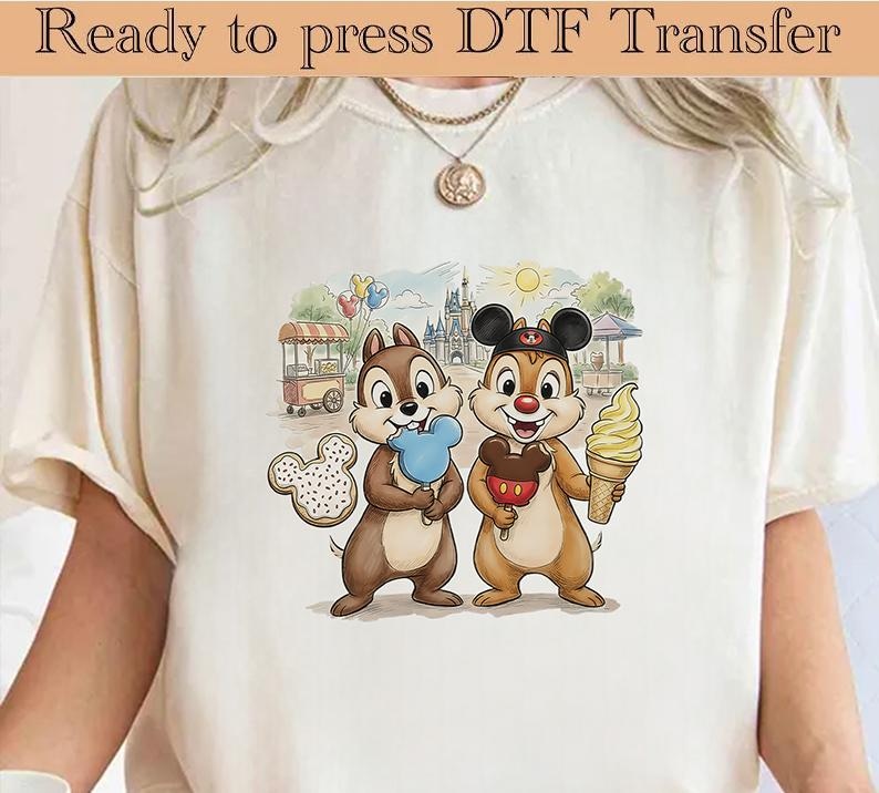 May include: Cream-coloured t-shirt with a DTF transfer of Chip and Dale. Chip holds a blue popsicle, and Dale holds a Mickey Mouse ice cream. The background shows a castle, food cart, and balloons.