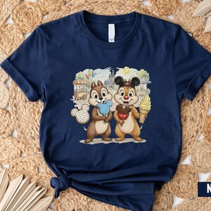 May include: Navy blue t-shirt with a colourful graphic of Chip and Dale holding treats in front of a theme park backdrop. The cartoon characters are holding a Mickey Mouse-shaped cookie, a red apple, and an ice cream cone.