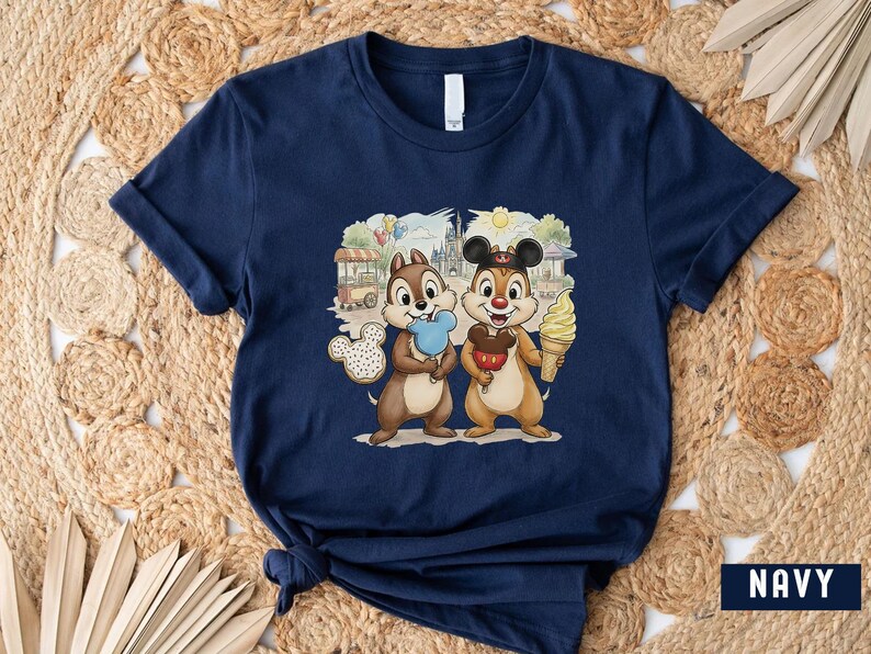 May include: Navy blue t-shirt with a colourful graphic of Chip and Dale holding treats in front of a theme park backdrop. The cartoon characters are holding a Mickey Mouse-shaped cookie, a red apple, and an ice cream cone.