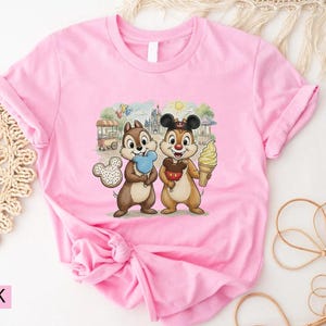 May include: Pink t-shirt with a graphic of Chip and Dale, holding treats in front of a theme park scene. Chip holds blue candy floss, and Dale holds an ice cream cone. The shirt has rolled-up sleeves.