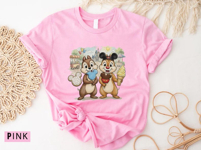 May include: Pink t-shirt with a graphic of Chip and Dale, holding treats in front of a theme park scene. Chip holds blue candy floss, and Dale holds an ice cream cone. The shirt has rolled-up sleeves.