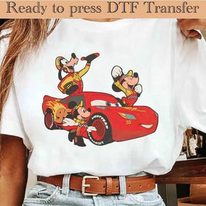Può includere: Una t-shirt bianca con una stampa grafica di un'auto da corsa rossa con il numero 95, accompagnata dai personaggi dei cartoni animati Pippo e Topolino in tenuta da gara. Il testo Ready to press DTF Transfer è stampato in alto nell'immagine.