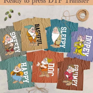 Puede incluir: Una colección de camisetas con los siete enanitos de Blancanieves. Cada camiseta es de un color diferente y tiene un personaje enano con su nombre impreso verticalmente. El texto "Ready to press DTF Transfer" está en la parte superior.