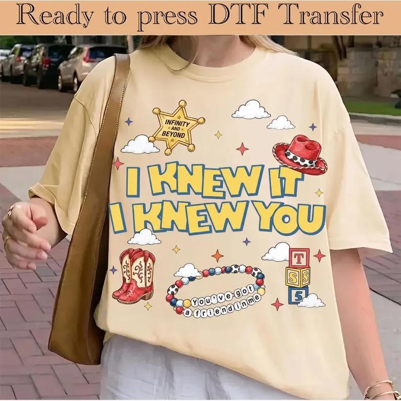 Pu&ograve; includere: Una t-shirt a maniche corte color crema con una stampa grafica che include una stella da sceriffo gialla, un cappello da cowboy rosso, stivali da cowboy rossi, un braccialetto di perline e blocchi impilati. Il testo recita I KNEW IT I KNEW YOU, INFINITY AND BEYOND e YOU'VE GOT A FRIEND IN ME.
