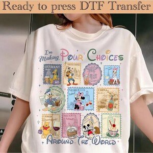 May include: Cream-colored t-shirt with a colorful graphic featuring Disney characters and stamps representing different countries. The text "I'm Making Pour Choices Around The World" is also visible.