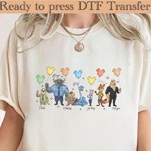 May include: Cream-colored t-shirt featuring a DTF transfer of Zootopia characters holding Mickey Mouse-shaped balloons. The characters include Flash, Bogo, Nibbles, Gary, Judy Hopps, Nick Wilde, and Mayor Lionheart. Text at the top reads "Ready to press DTF Transfer."