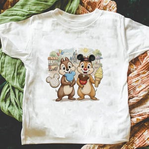 May include: White t-shirt with a colourful illustration of Chip and Dale, Disney characters, enjoying treats in front of a park scene. One holds blue candy floss, the other an ice cream cone. A Mickey Mouse-shaped biscuit is also visible.