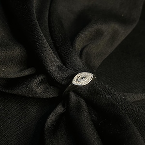 May include: A silver ring featuring a marquise-shaped setting, adorned with small, clear gemstones. The ring is displayed on a dark, textured fabric, which accentuates the ring's design. The ring has a slender, delicate band.