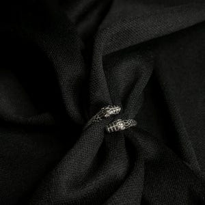 Könnte beinhalten: Ein silberner Schlangenring mit detaillierten Schuppen und zwei sich gegenüberliegenden Schlangenköpfen. Der Ring ist vor einem dunklen, strukturierten Stoffhintergrund platziert, was einen starken Kontrast erzeugt.