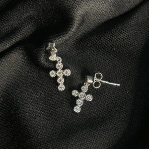 Puede incluir: Un par de pendientes de cruz plateados con piedras preciosas redondas y transparentes. Los pendientes se muestran sobre un fondo negro texturizado. El diseño de cruz es una elección de joyería clásica.