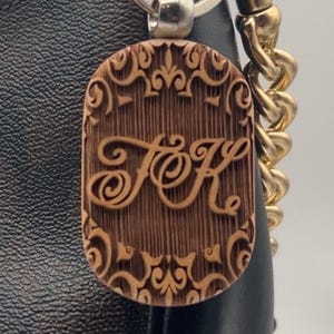 May include: A brown wooden keychain with the initials "FK" in an ornate font, attached to a black and brown Coach handbag. The keychain has a decorative border and is connected to a silver keyring and gold chain.