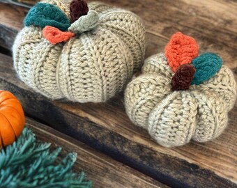 May include: Two beige crocheted pumpkins with colorful leaf accents. One pumpkin is larger than the other. The pumpkins are on a wooden surface with small orange pumpkins and green foliage scattered around.